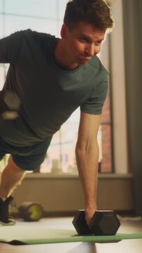 Vertical Screen: Fit Young Man Performing Enduring Training in Plank Position, while Lifting Dumbbells During Morning Workout at Home in Sunny Apartment. Concept of Healthy Lifestyle and Fitness.