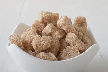 Cube-shaped cane sugar in porcelain bowl on white wooden table. Front closeup view, no people.