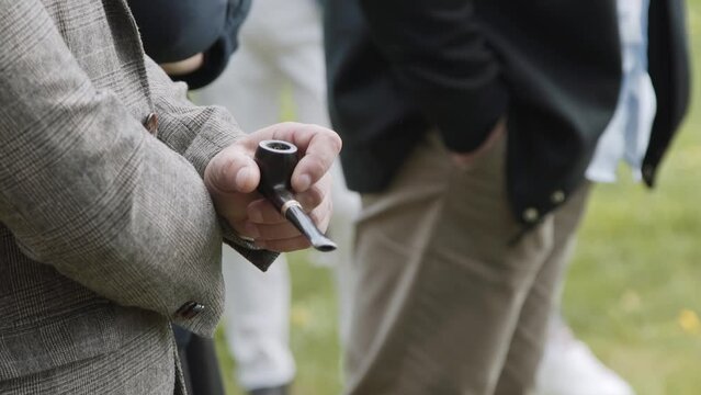 Man in business suit, in company of other men, stands on green lawn and holds wooden smoking pipe in his hand.