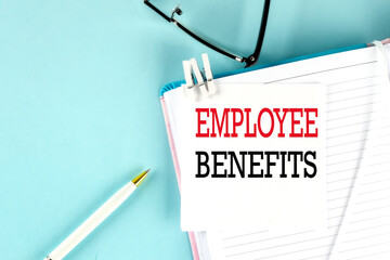 EMPLOYEE BENEFITS text on a sticky on notebook with pen and glasses , blue background