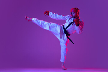 Studio shot of male taekwondo fighter in sports uniform in action isolated over purple background...