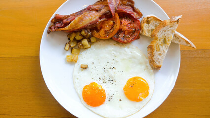 American breakfast with sunny side fried eggs, bacon, tomatoes, toasted bread and baked potatoes on wooden table in restaurant. Delicious served lunch or brunch food set meal in cafe. Top view.