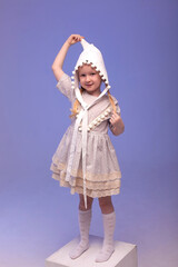 Cute little girl with pigtails in a beautiful dress and cap dancing on a light blue background. He smiles and looks at the camera. Space for text