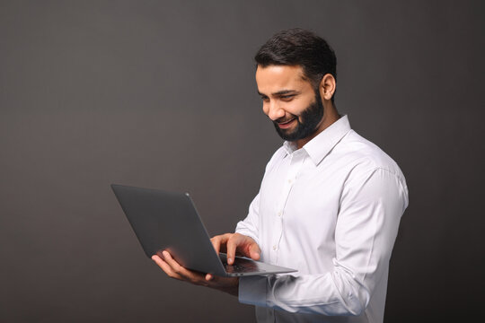 Portrait of happy young bearded Indian man in formal white shirt holding and using laptop computer isolated on black background. Male office employee typing, answering email, copy space