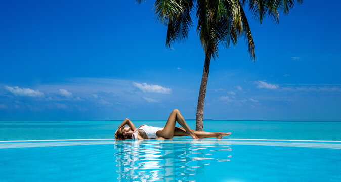 Elegant tanned woman in white swimsuit in pool on tropical Maldives island. Beautiful bikini body girl in pool with view on horizon. Sexy model near the pool on beautiful Indian ocean landscape. 