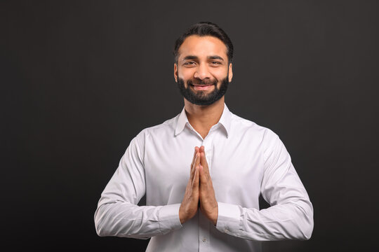 Happy bearded Indian man in formal white shirt greeting or thanking you with traditional gesture namaste standing isolated on black background. Male office employee welcoming, holding arms in pray