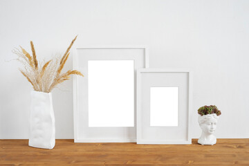 Mockup of two white photo frames on a wooden table with a vase of dried flowers and a plaster bust.