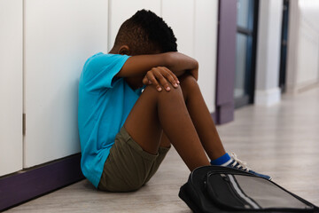 Sad african american elementary schoolboy hugging knees while sitting on floor in corridor
