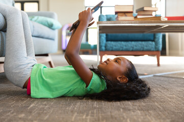 Side view of african american elementary girl using digital tablet while lying on carpet in school