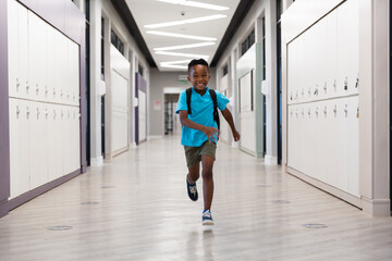 Full length of smiling african american elementary schoolboy running in corridor at school - Powered by Adobe
