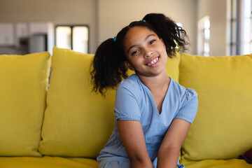 Portrait of smiling biracial elementary schoolgirl with head cocked sitting on couch in school
