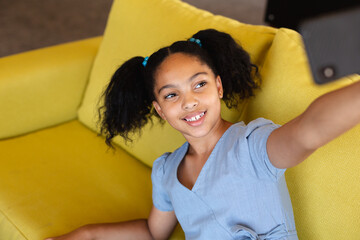 Smiling biracial elementary schoolgirl taking selfie while sitting on couch in school