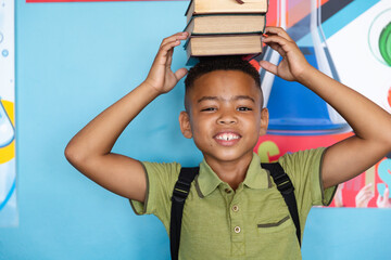 Portrait of smiling african american elementary schoolboy stacking books on head against wall