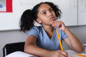 Thoughtful biracial elementary girl with hand on chin looking up while studying at desk in classroom - Powered by Adobe