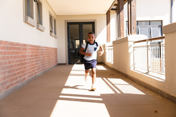 Full length of african american elementary schoolboy with backpack running in school corridor