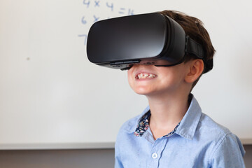 Smiling caucasian elementary schoolboy wearing vr glasses against whiteboard in classroom