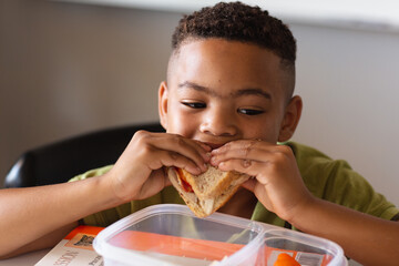 Close-up of african american elementary schoolboy eating sandwich at desk during lunch break - Powered by Adobe