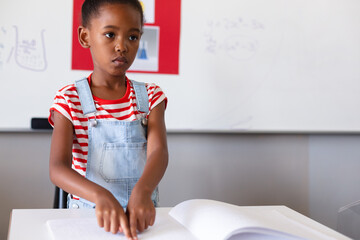 Blind african american elementary schoolgirl touching braille book while studying in classroom - obrazy, fototapety, plakaty