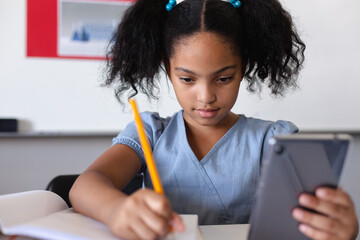 Biracial elementary schoolgirl using digital tablet while writing on book at desk in classroom