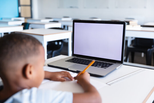 African american elementary schoolboy writing on book while using laptop at desk in class