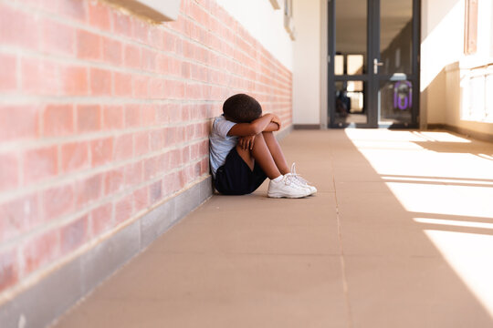 Full length of sad african american elementary schoolboy sitting on floor in school corridor