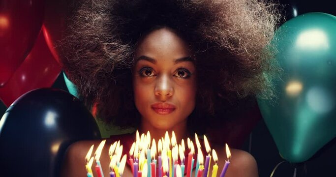 Young woman at her birthday party celebrating, holding a cake with candles. Young African American woman with an afro holding her birthday cake looking at the candles surrounded by colourful balloons