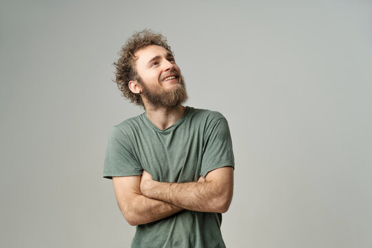 Smiling handsome young man with bearded and wild curly hair, bright blue eyes looking up with hands folded isolated on white background. Young thinking man in green t-shirt on white background