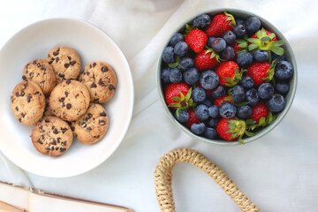 Picninc blanket with straw bag, bowl of strawberries and blueberries, bowl of chocolate chip cookies, books, sunglasses and basket of apples in the garden. Top view.