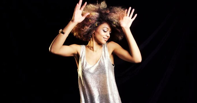 Carefree african american woman dancing against a black background with gold confetti in her hair. Young woman having fun at a party celebrating and dancing against a dark background