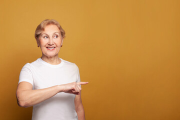 Happy smiling mature woman pointing at copy space on bright yellow studio wall background