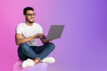 Full length of millennial Arab man in glasses using laptop for work or studies in neon light, copy space