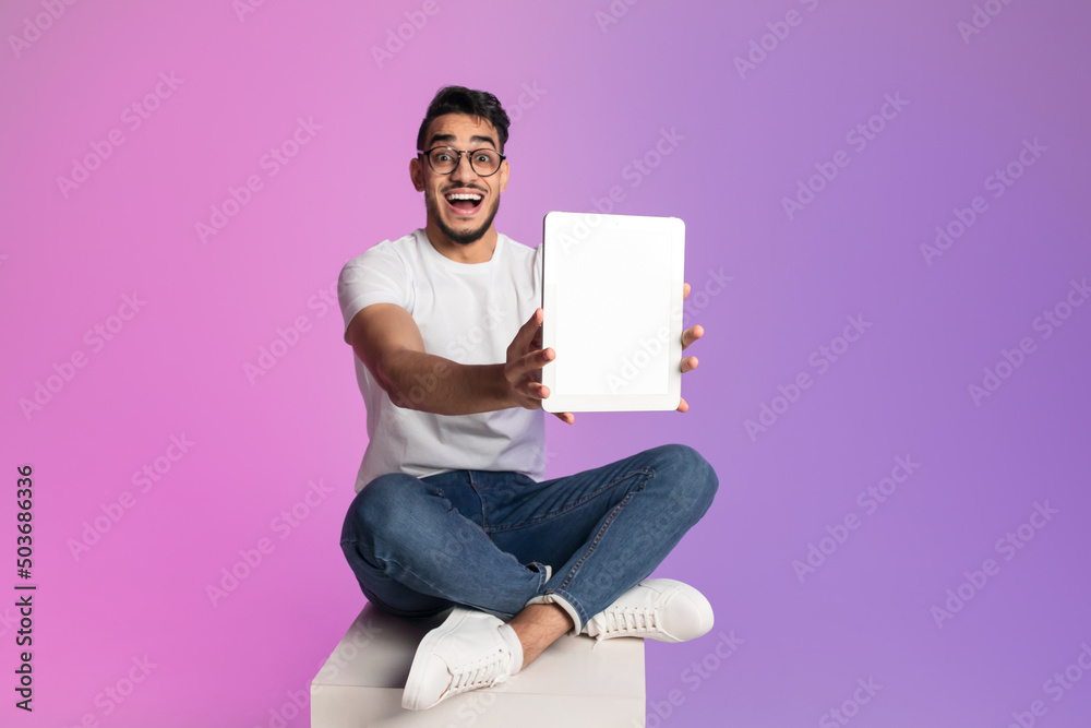 Poster excited millennial arab man showing tablet with blank screen in neon light, mockup for app or websit