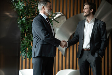 Two smiling businessmen shaking hands together while standing by windows in an office boardroom.