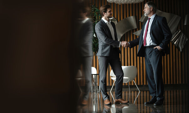 Two smiling businessmen shaking hands together while standing by windows in an office boardroom.