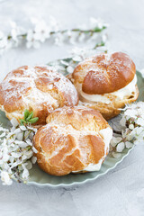 Choux Buns with whipped cream and sugar powder on top on plate. Gray background with spring white cherry blossom flowers. Delicate choux pastry dessert. French cream puffs, close-up