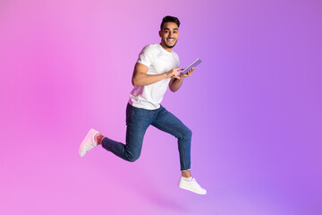 Happy young Arab man with smartphone jumping up in neon light, copy space. Cool new app, website or advertisement