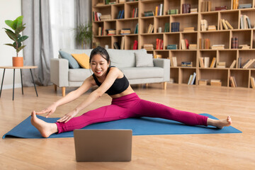 Home workout videos. Sporty asian lady in sportswear stretching on yoga mat while watching online tutorial on laptop