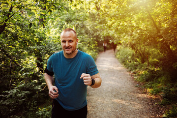 Smiling male with in-ear headphones jogging in park. Mid adult man is listening to music during summer. He is wearing sports clothing. Shot of a young man going for a morning run.