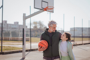 Happy father and teen daughter embracing and looking at camera outside at basketball court.