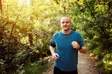 Smiling male with in-ear headphones jogging in park. Mid adult man is listening to music during...