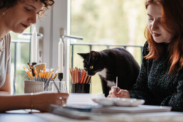 Two Caucasian women painting together, a cat next to them - Powered by Adobe