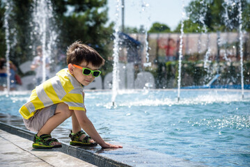 Little boy plays in the square near pool with water jets in the fountain at sunny summer day. Active summer leisure for kids in the city