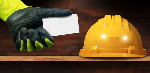 Closeup of a yellow Safety Helmet above a wooden workbench and a Manual worker with green and black...