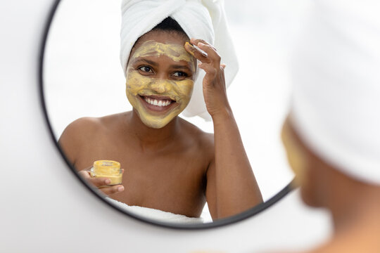 Smiling african american mid adult woman looking in mirror and applying face pack
