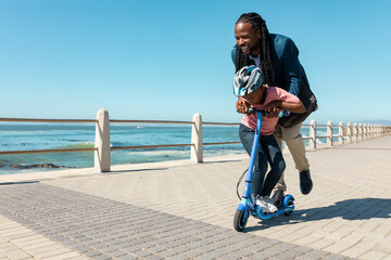 Full length of happy african american father and son riding push scooter together at promenade