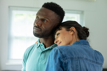 Multiracial young couple looking away while embracing in living room at home