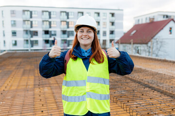 Architect doing ok sign over building background, thumb up with fingers. Portrait of successful woman constructor wearing white helmet and safety yellow vest. Woman are planning new project.