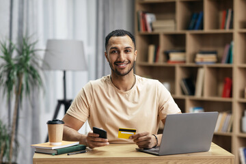 Obraz premium Cheerful arab man purchasing goods on web using smartphone, laptop and credit card, enjoying shopping in internet