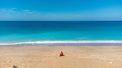 Fototapeta na wymiar Aerial view of woman in red dress, sitting on the sandy beach, enjoying soft turquoise ocean wave. Tropical sea in summer season on Megali Petra beach on Lefkada island.