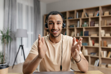 Positive arab freelancer guy in headphones having online video call, speaking to business partner and gesturing at camera